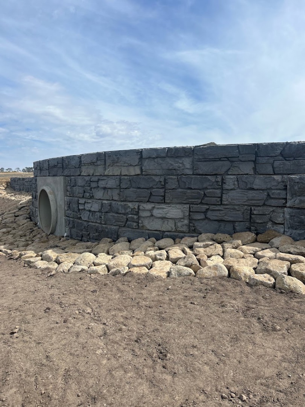 A curved rock-faced block retaining wall with a culvert outlet and rock beaching