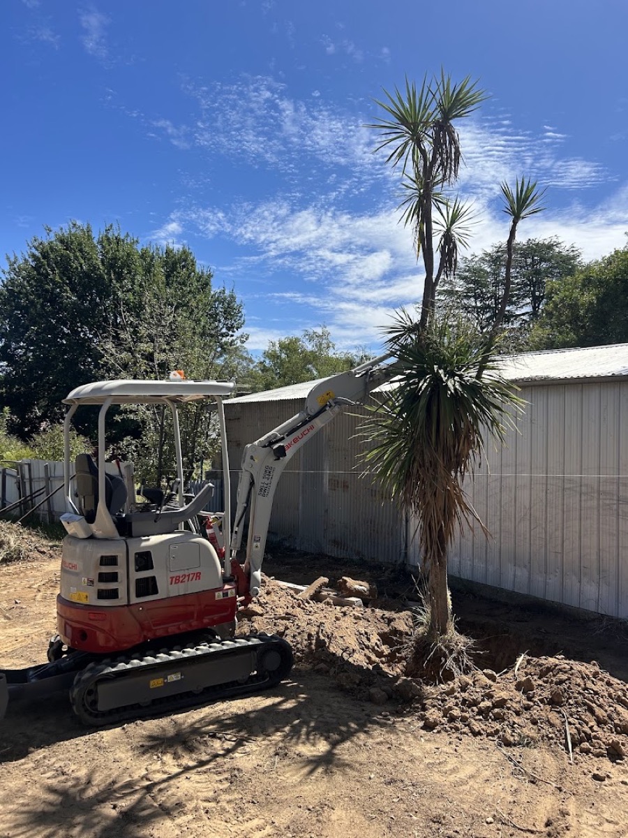 A mini excavator working on stripped ground beside a shed