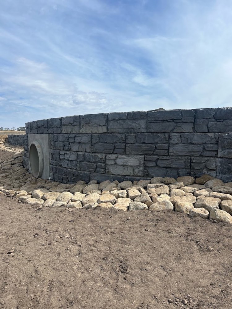 A curved rock-faced retaining wall with a culvert outlet and rock beaching at its base