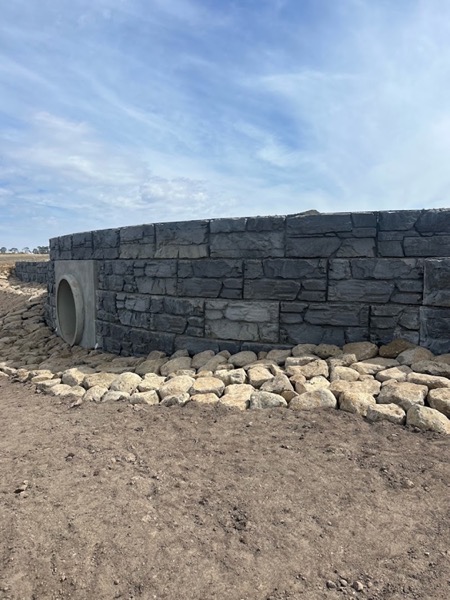 A curved rock-faced retaining wall with a culvert outlet and rock beaching at its base