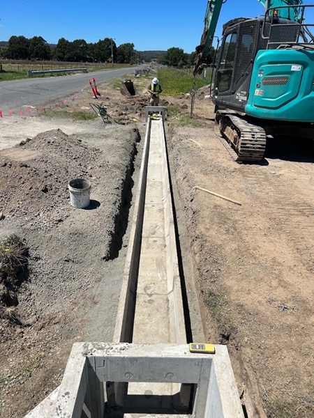 An excavator beside an open roadside trench with a precast channel bedded in