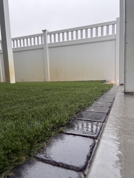 A close view of synthetic turf meeting a charcoal paver edge and wet concrete